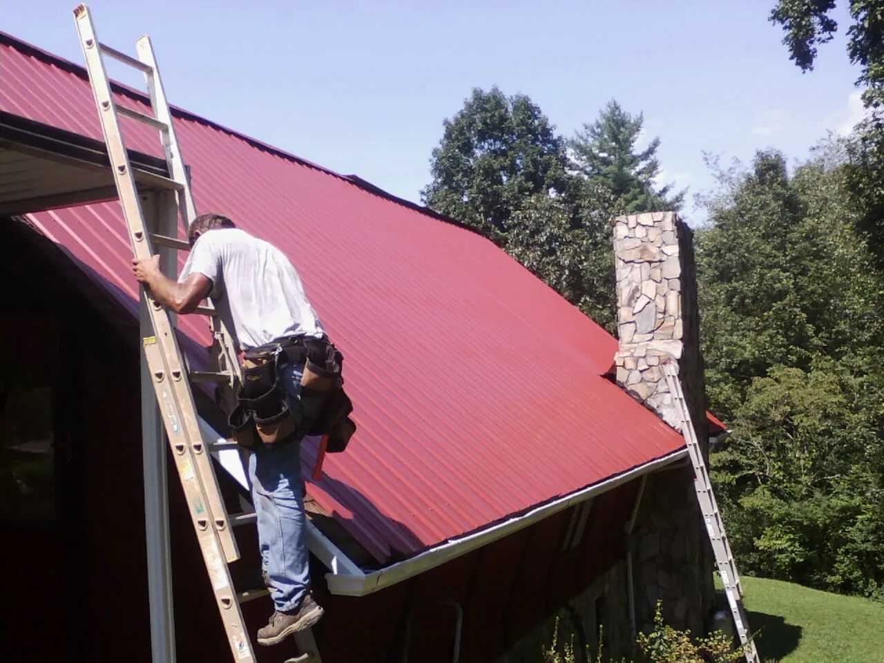Roofer on a ladder beside a red metal roof and stone chimney — Soffit & Fascia Repair by Roofing Brothers in Forest Hill